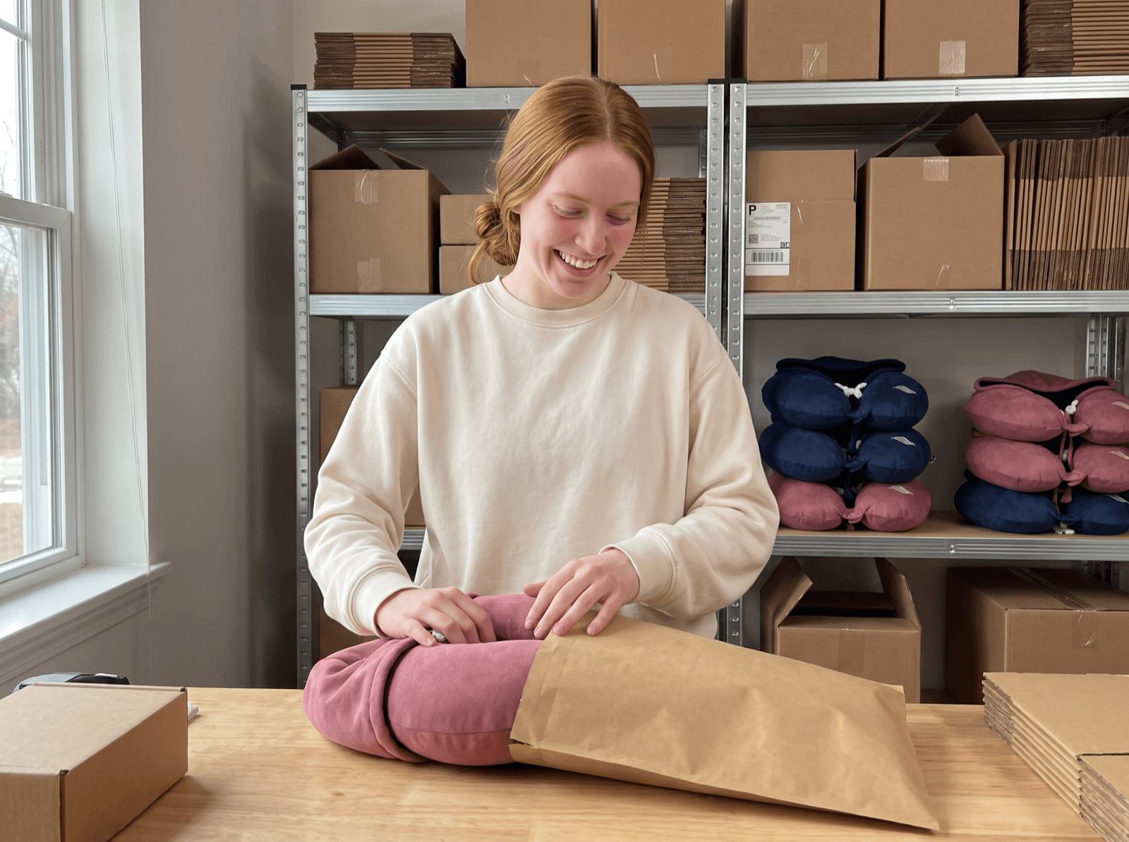 Rikke packing a dusty rose Snug Hood into a shipping mailer at the warehouse table.